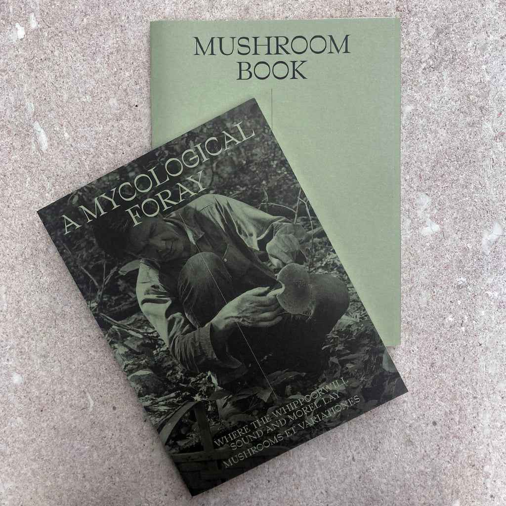 Photo of the two books contained within the packaging: A MYCOLOGICAL FORAY printed with a full-bleed cover photo of John Cage foraging in black and white printed on green paper, and MUSHROOM BOOK, a plain green envelope labled only with its title.
