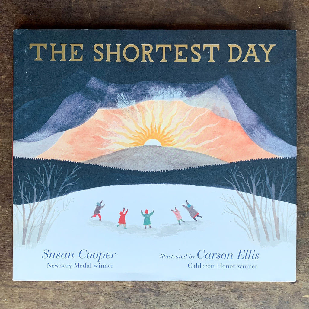 Hard front cover of The Shortest Day with an illustration of folks frolicking in the snow while the sunsets over a hill and darkness falls like a curtain.