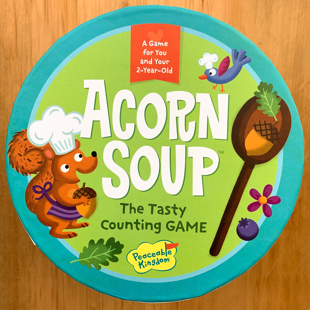 Top of game box, circular with tect "Acorn Soup, The Tasty Counting GAME" with illustrations of a squirrel and bird in chefs hats and assorted woodland objects