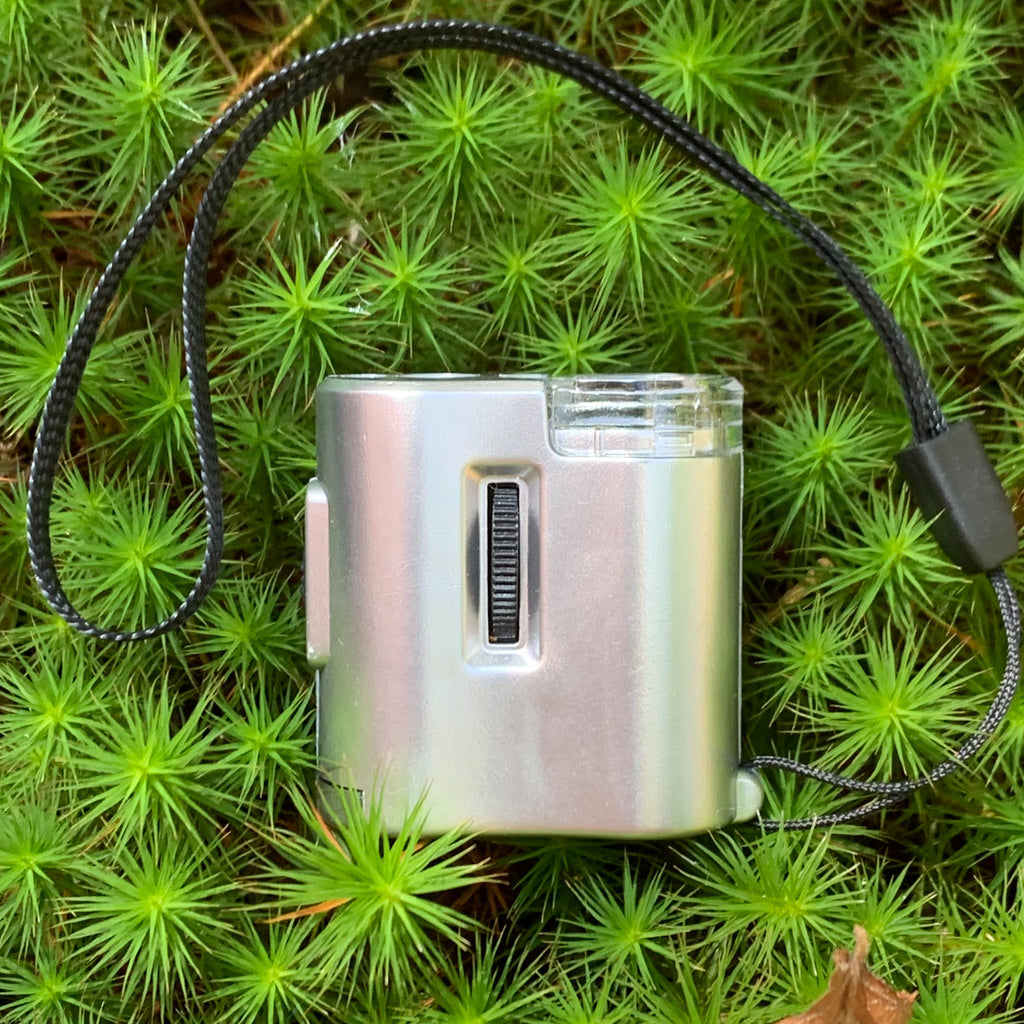Silver plastic pocket microscope with lanyard against a verdant backdrop of small plants.