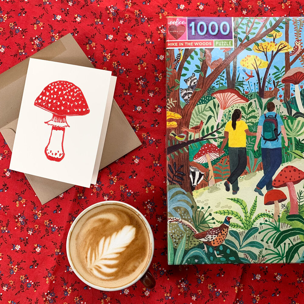 Greeting card with red block printed amanita mushroom on a white background shown on a table next to a jigsaw puzzle box and a latte.