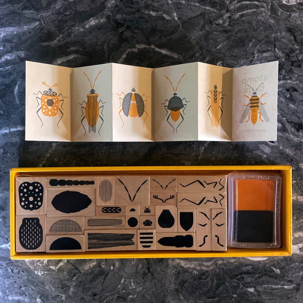 Inside contents of Stamp Bugs box including 25 wooden-backed rubber stamps, a black inkpad, a yellow inkpad, and a small booklet describing the kit.