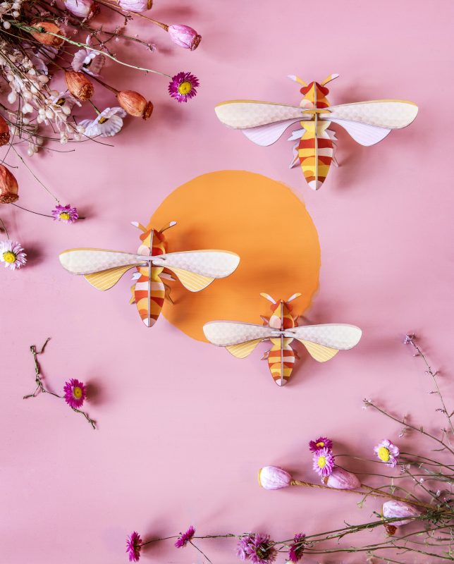 3 pink, red, and yellow stylized honeybees on a pink wall backdrop with dried flowers 