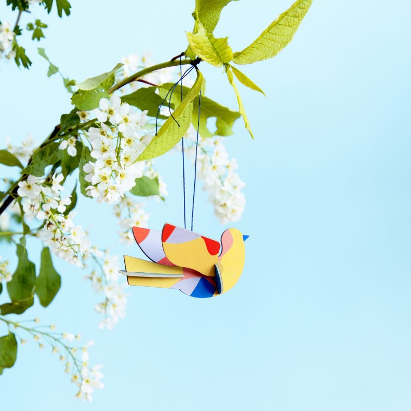 Yellow and color-block dove ornament hanging on a blossoming white shrub against a blue backdrop