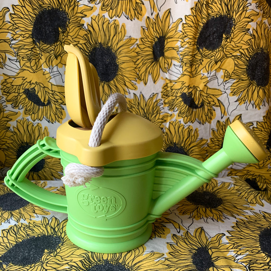 Green recycled plastic watering can with bellow accents and a rope handle against a backdrop of sunflower printed fabric.